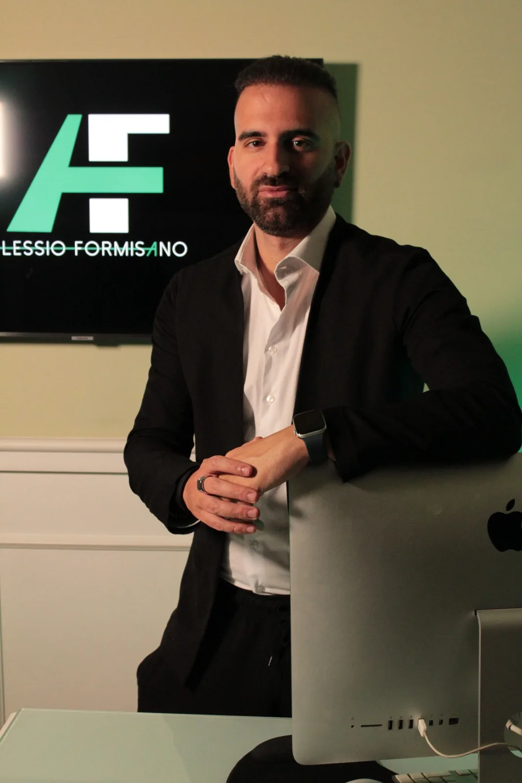 Alessio Formisano in piedi nello studio, accanto al logo